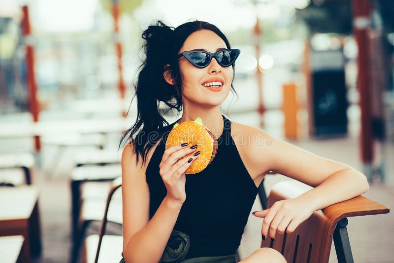 Young beautiful woman eating burger and enjoying while sitting outdoor. Fastfood Unhealthy Lifestyle Fashion Food concepts