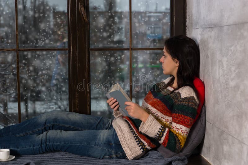 Young beautiful woman holding a book, lying on the windowsill an