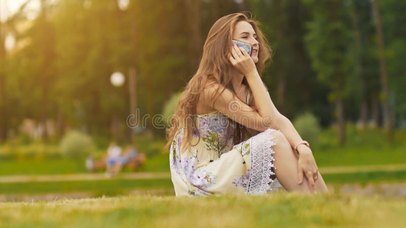Young beautiful woman in summer dress with long hair sitting on grass in green park and talking on the phone, smiling stock photography Young beautiful woman in summer dress with long hair sitting on grass in green park and talking on the phone, smiling stock photography