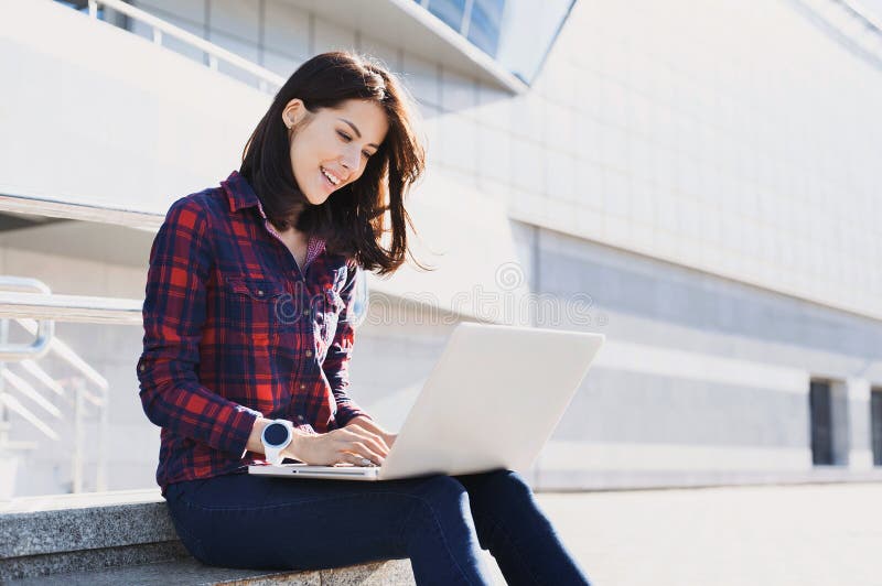 Young beautiful woman using laptop computer in the city street. Pretty happy girl working on laptop outdoor.