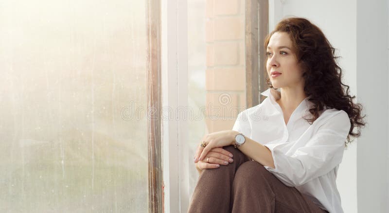 A young beautiful woman in a white shirt sits with a pensive view on the windowsill and looks out the window. The