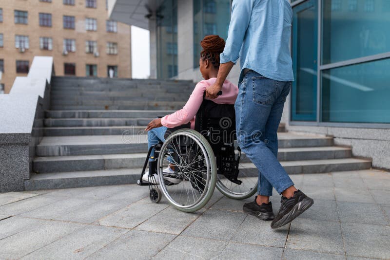 Black guy pushing wheelchair with handicapped woman, having problem entering building, standing near stairs without ramp