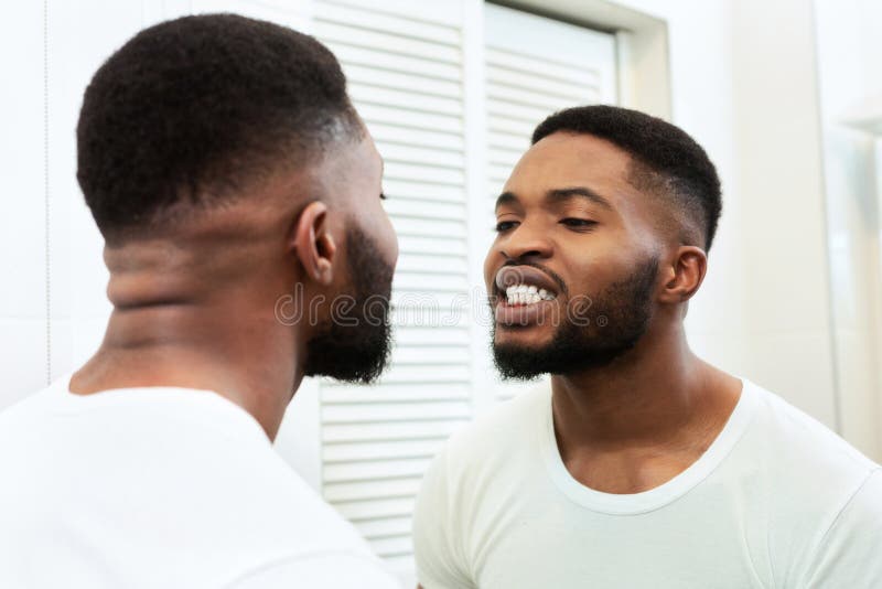 Young black man looking in mirror at his teeth in bathroom