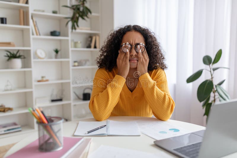 Young black woman in glasses rubbing irritated eyes, sitting at desk with laptop, studying online too much at home royalty free stock photography
