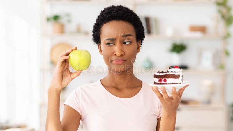 Young black woman looking at apple with suspicion, not wanting to eat fruit instead of cake at home