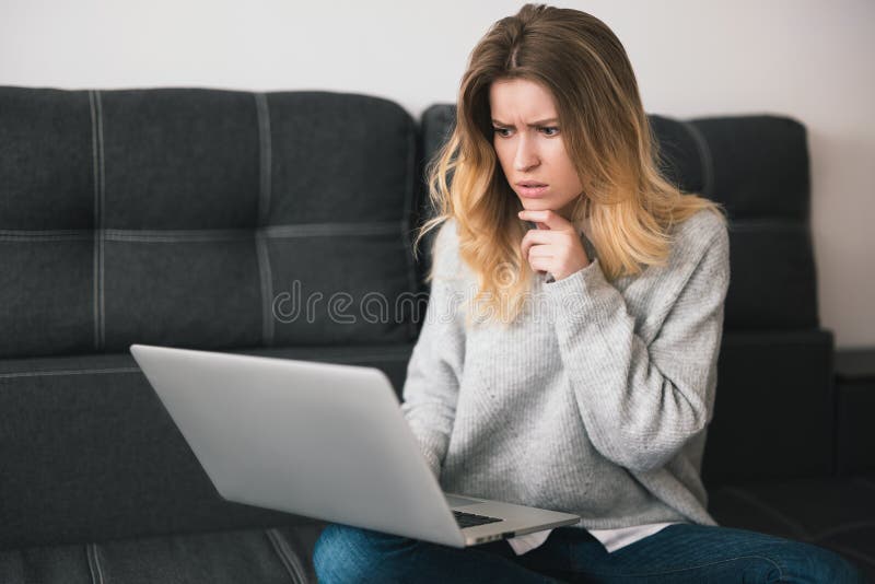 Young blond beautiful woman sits on the sofa working from home on her laptop during quarantine looks thoughtful, cozy home