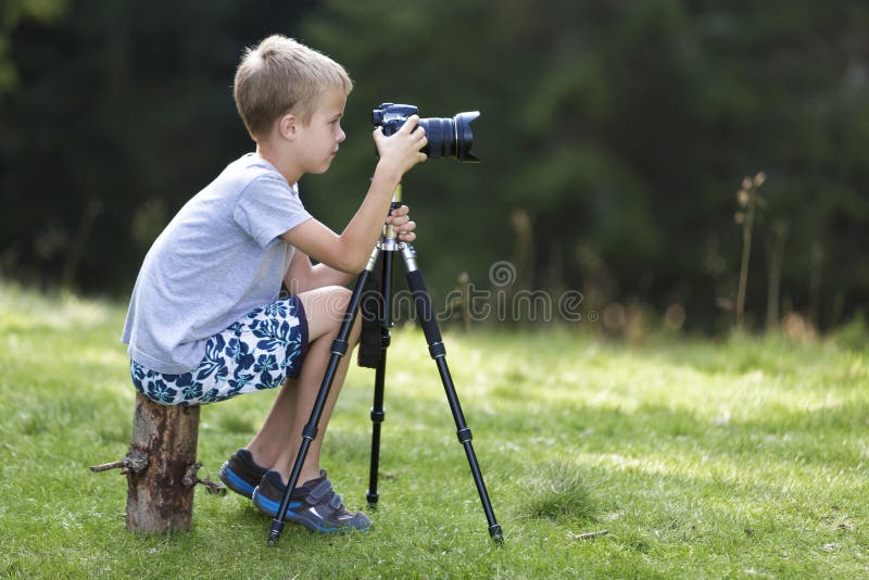 Young blond child boy sitting on tree stump on grassy clearing taking picture with tripod camera