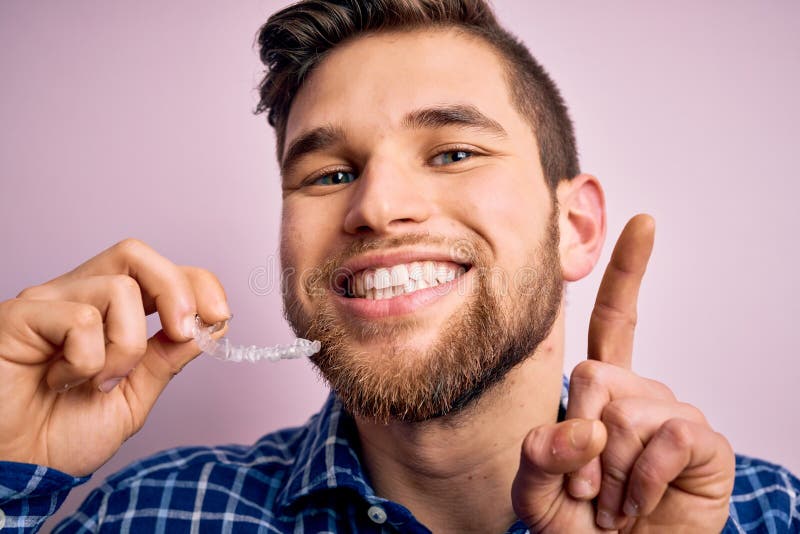 Young blond man with beard and blue eyes holding dental aligner over pink background surprised with an idea or question pointing