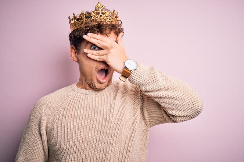 Young blond man with curly hair wearing golden crown of king over pink background peeking in shock covering face and eyes with royalty free stock photo