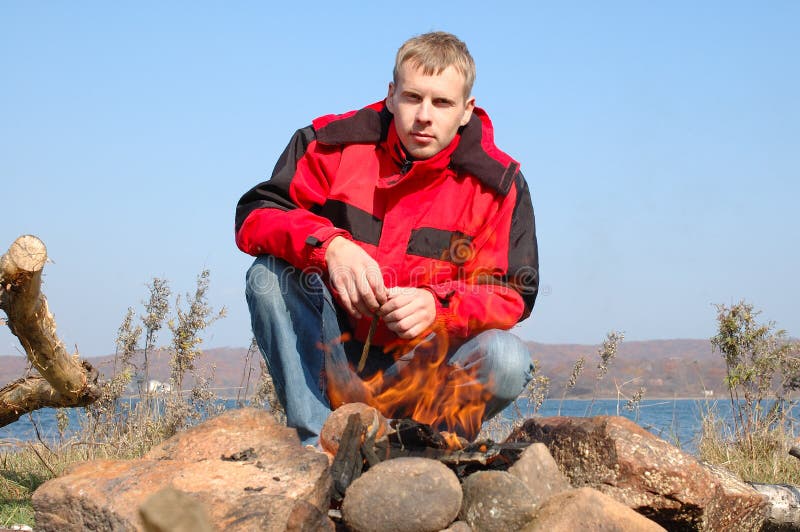 Young blond man in red jacket sit near fire.