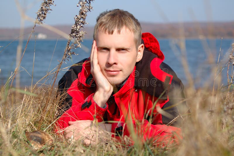 Young blond man in red jacket sit on the seashore.