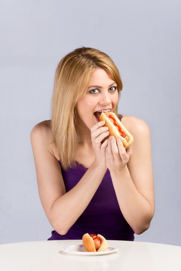 Beautiful blonde female biting hot dog bun with sausage and ketchup during lunch break. The second helping is in front of her on table placed paper plate. Beautiful blonde female biting hot dog bun with sausage and ketchup during lunch break. The second helping is in front of her on table placed paper plate.