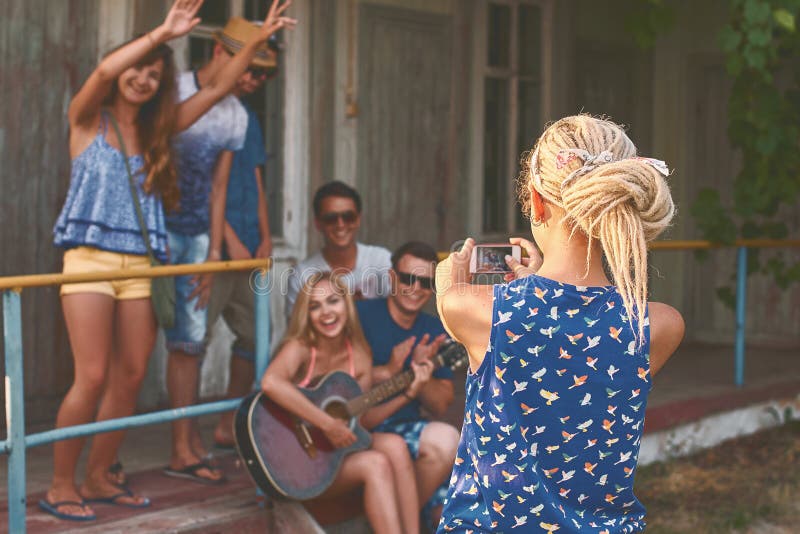 Young blonde girl with dreads takes a photo of a groups of her friends with her smartphone near the wooden holiday cabin stock photos Young blonde girl with dreads takes a photo of a groups of her friends with her smartphone near the wooden holiday cabin stock photos
