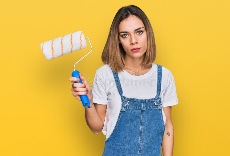 Young blonde girl holding roller painter thinking attitude and sober expression looking self confident