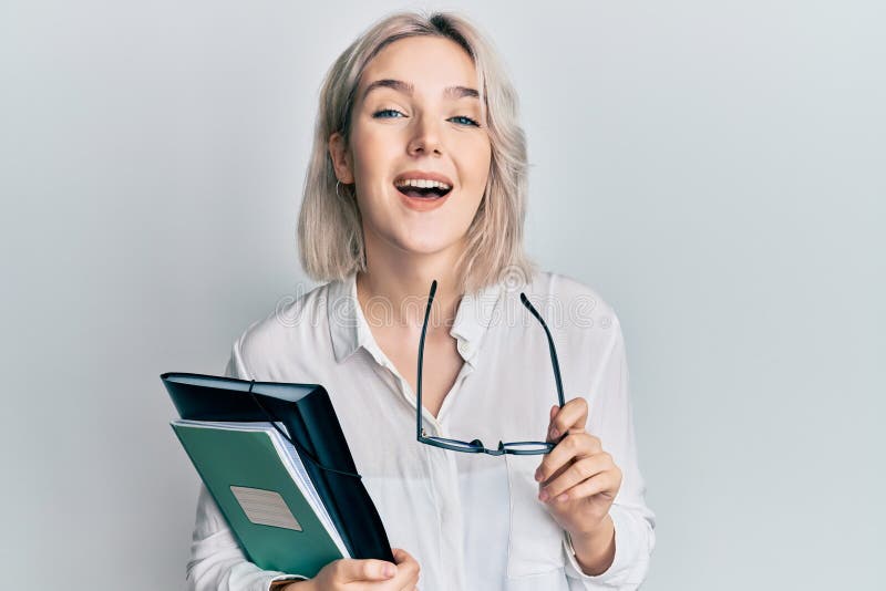 Young blonde girl reading a book and holding glasses smiling and laughing hard out loud because funny crazy joke