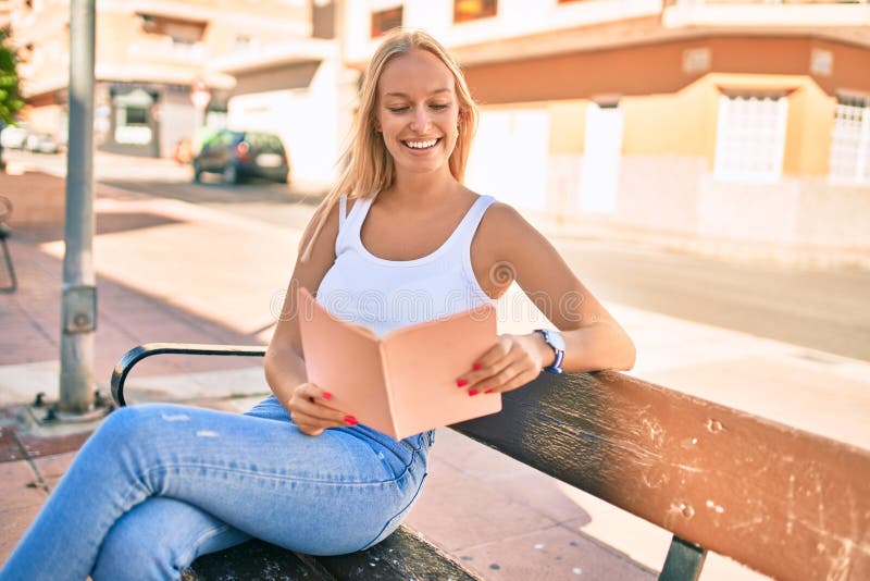 Young blonde girl smiling happy reading book sitting on the bench at the park