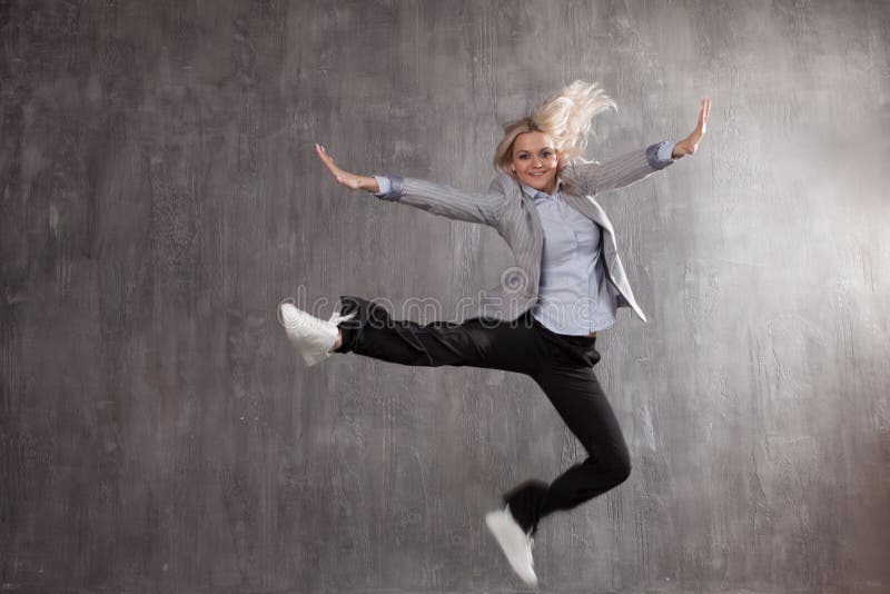 Young blonde woman in business suit and sneakers jumping for joy, gray textured background.