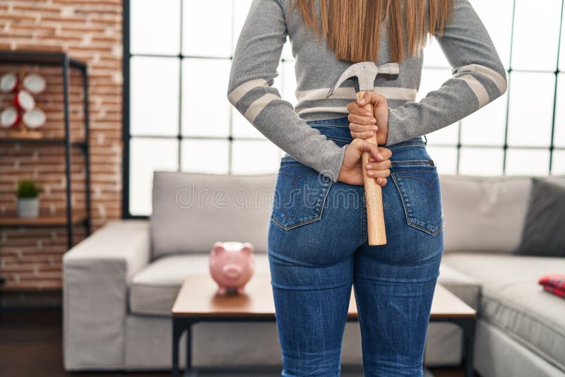 Young blonde woman holding hammer on back looking piggy bank at home