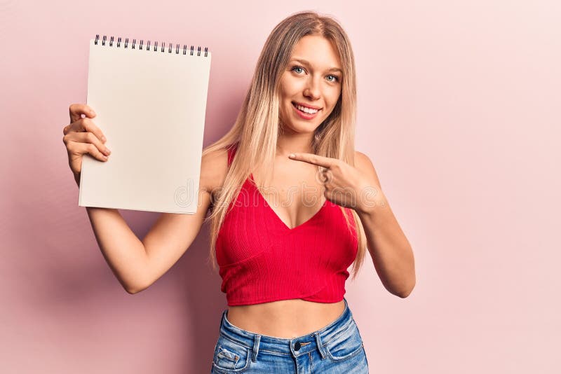 Young blonde woman holding notebook smiling happy pointing with hand and finger