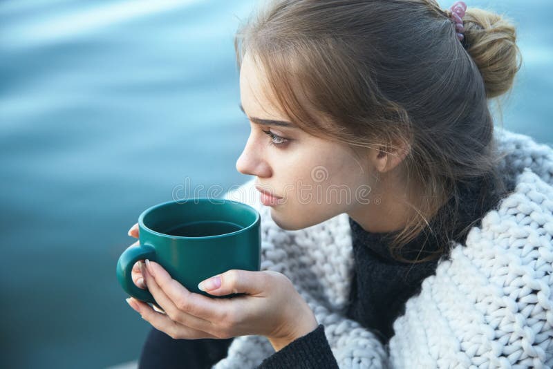 Young blonde woman wearing scarf holding coffee cup, warming up and drinking coffee or tea