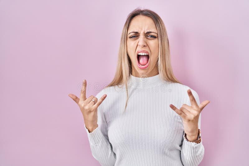 Young blonde woman wearing white sweater over pink background shouting with crazy expression doing rock symbol with hands up