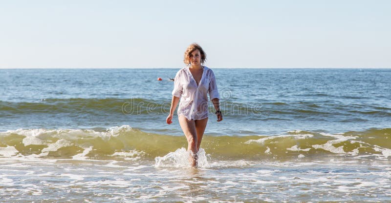 Young blonde woman in a wet white shirt comes out of the water near the seashore