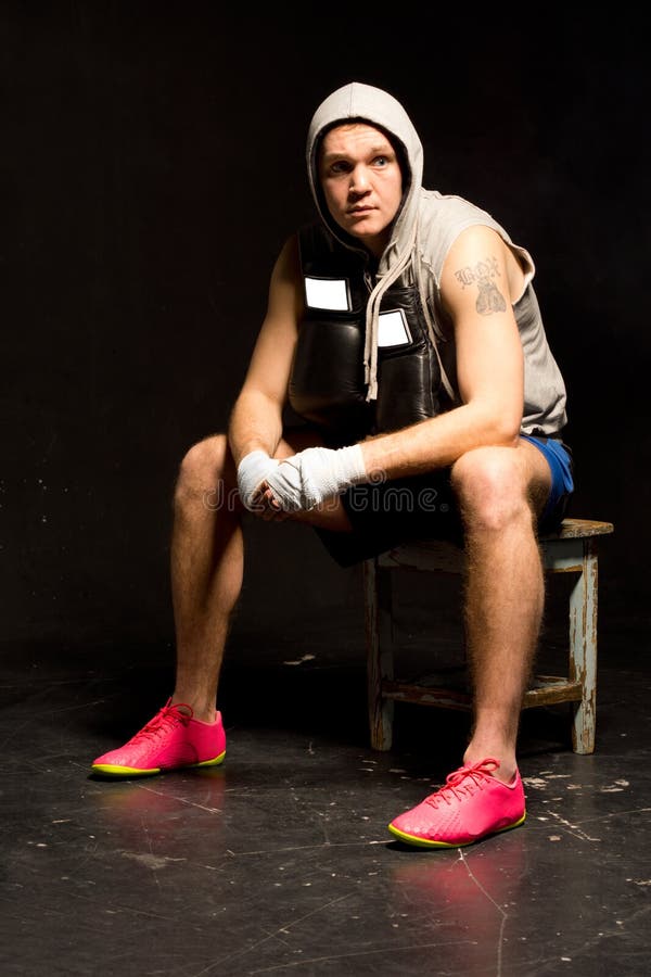 Young boxer taking a break from training sitting on a stool wearing a hoodie resting his bandaged fists on his knee staring thoughtfully to the side as he watches an opponent, dark background