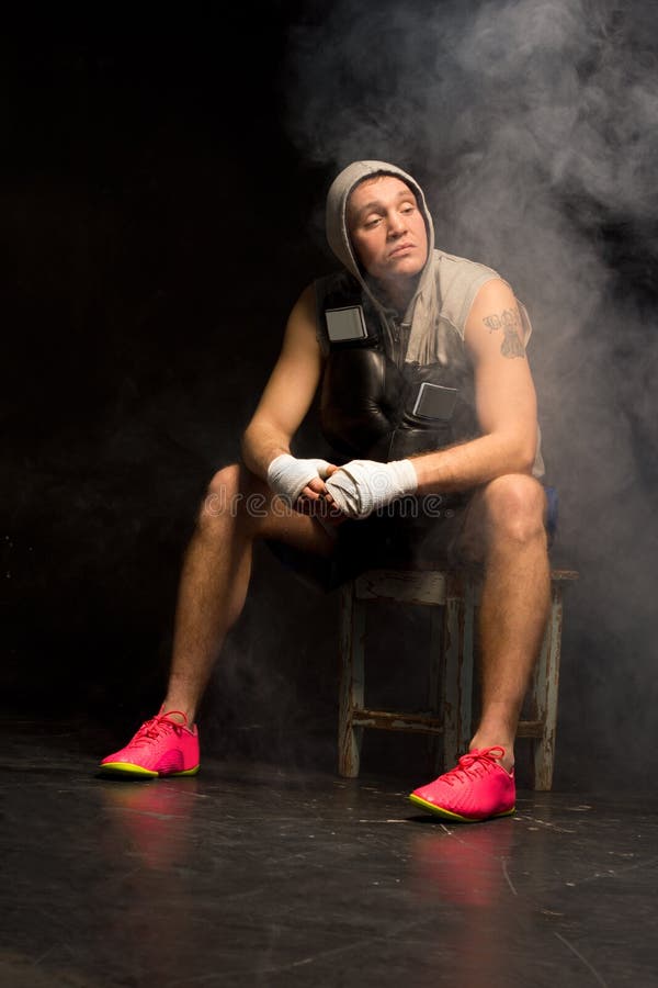 Young, boxer waiting patiently for his match sitting quietly on a wooden stool in a hoodie with his hands already bandaged and his gloves dangling around his neck