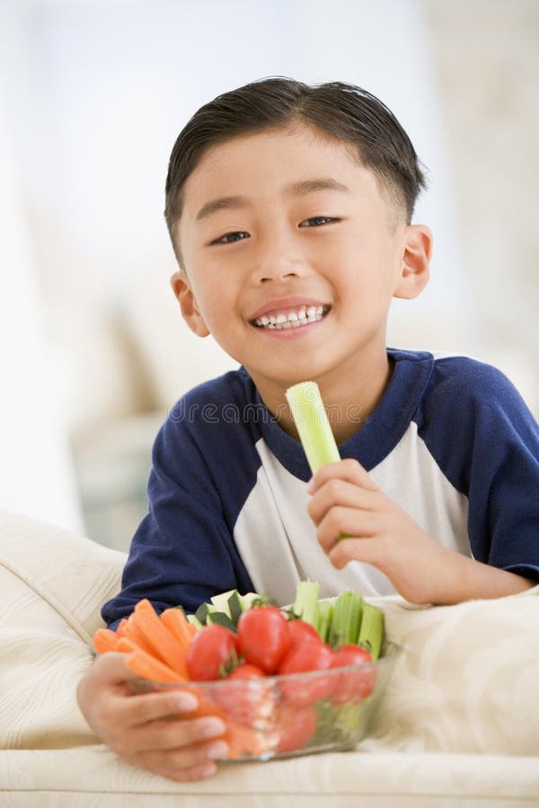 Mladý chlapec jesť misku zeleniny, v obývačke s úsmevom.