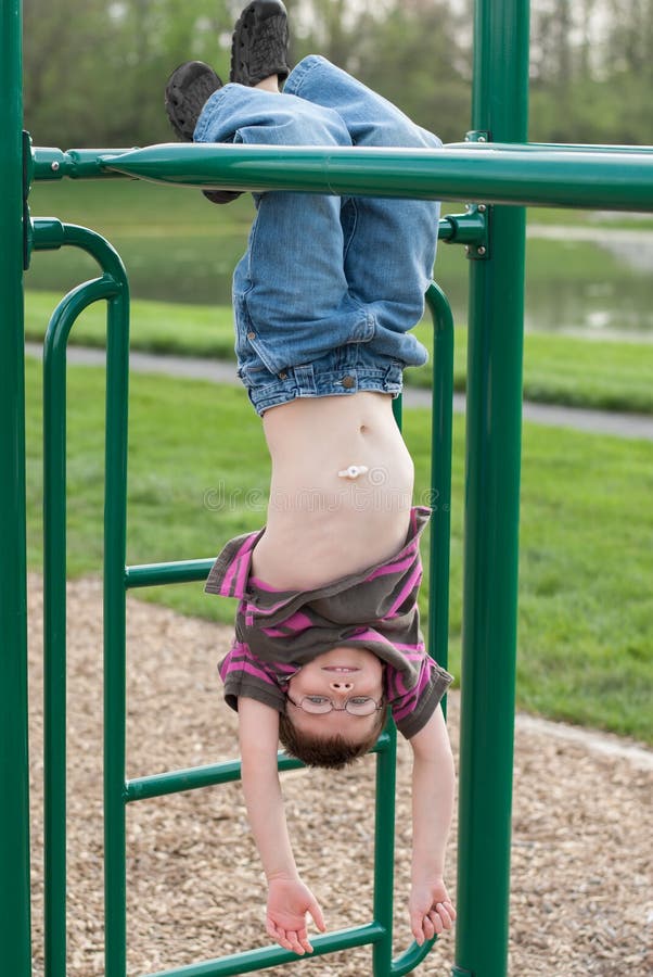 Young boy with gastric g-tube hanging upside down from monkey bars on jungl...