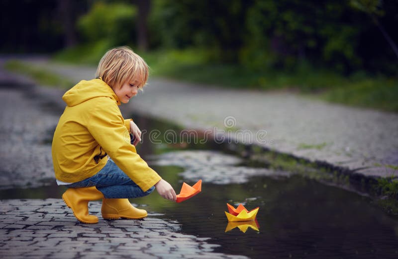 Young boy in rain boots and coat is putting paper boats on the water, at spring rainy day stock photography Young boy in rain boots and coat is putting paper boats on the water, at spring rainy day stock photography