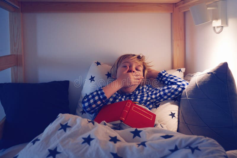 Young boy yawning while laying in the bed and reading a book before going to sleep