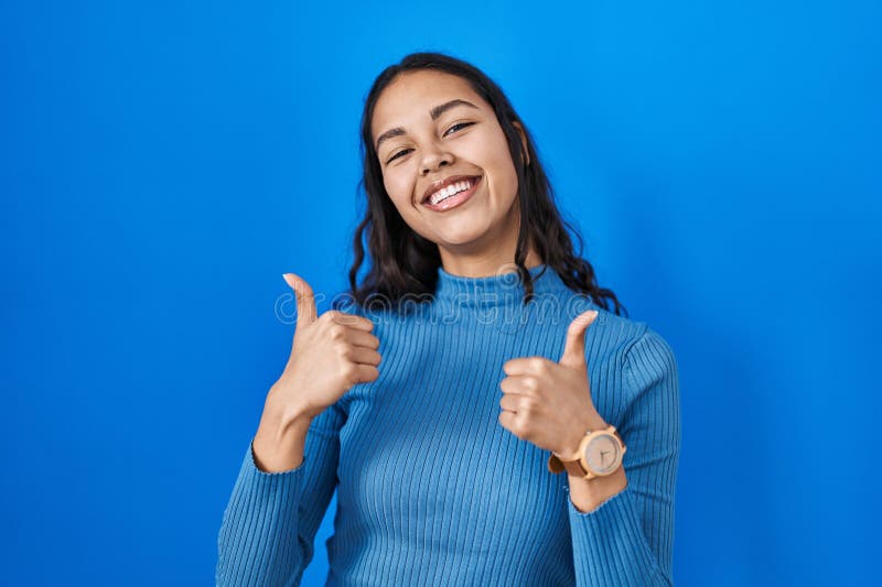 Young brazilian woman standing over blue isolated background success sign doing positive gesture with hand, thumbs up smiling and