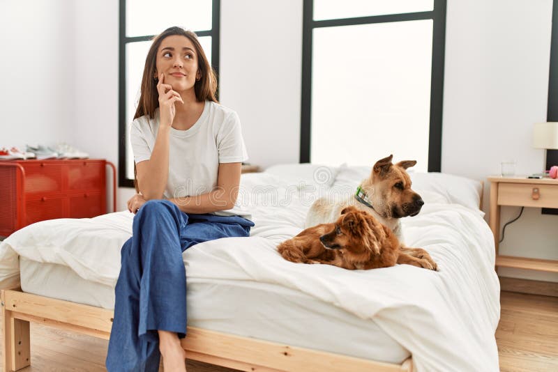 Young brunette woman sitting on the bed with two dogs serious face thinking about question with hand on chin, thoughtful about