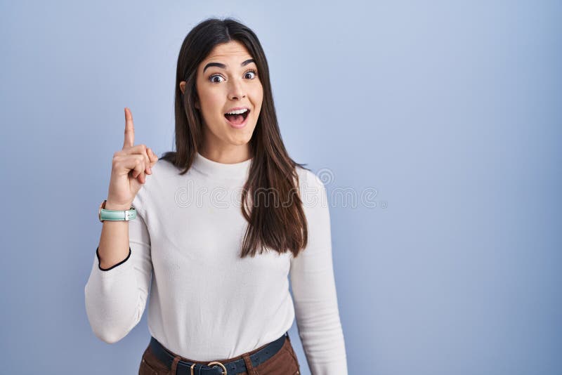 Young brunette woman standing over blue background pointing finger up with successful idea