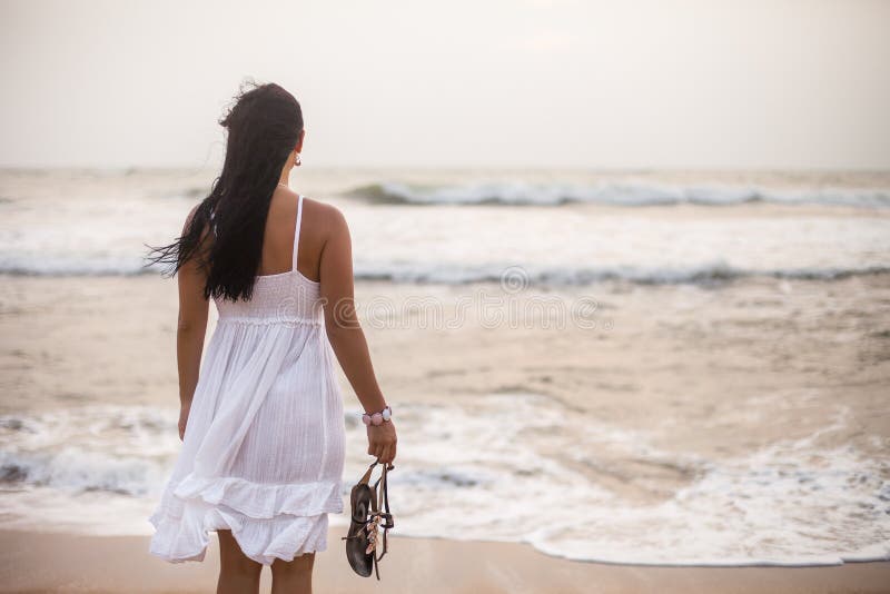 Young brunette woman in summer white dress standing on beach and looking to the sea. girl relaxing on vacation stock image Young brunette woman in summer white dress standing on beach and looking to the sea. girl relaxing on vacation stock image