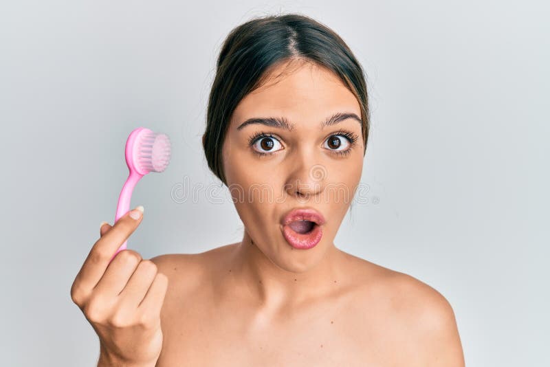 Young Brunette Woman Using Facial Exfoliating Brush Scared And Amazed With Open Mouth For