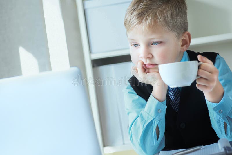 Young business boy working on laptop holding white cup of coffee or hot tea. Funny little boss in office.