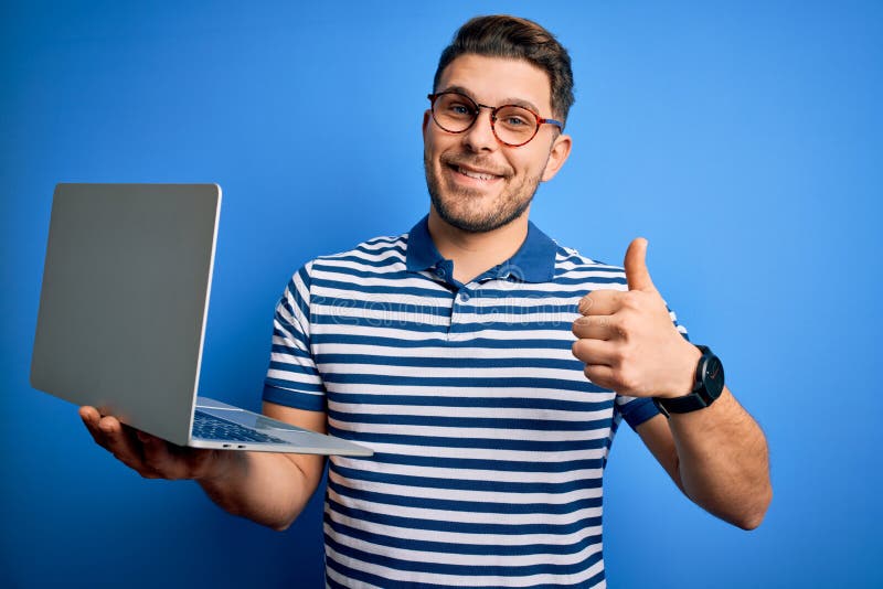 Young business man with blue eyes wearing glasses working using computer laptop happy with big smile doing ok sign, thumb up with fingers, excellent sign