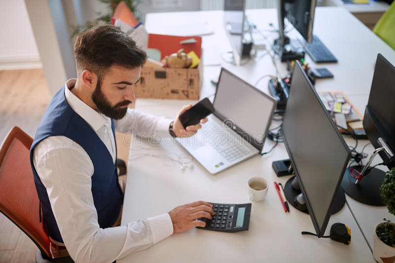 Young business man using calculator at his desk in office, processing data, holding cell phone in front of computer. business royalty free stock images