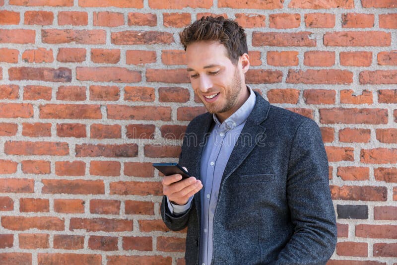 Young business man using smartphone in urban city background relaxing on brick wall texting sms on phone app living a