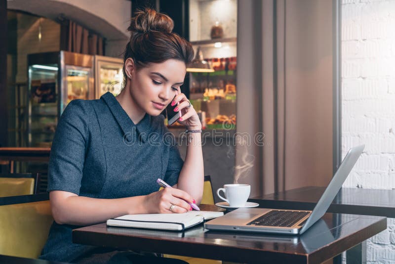 Young business woman in gray dress sitting at table in cafe, talking oncell phone while taking notes in notebook.