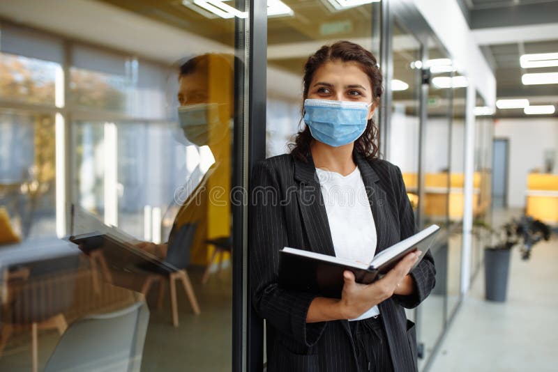 Young business woman with a notebook in her hands stands in the office corridor wearing medical mask to protect from coronavirus