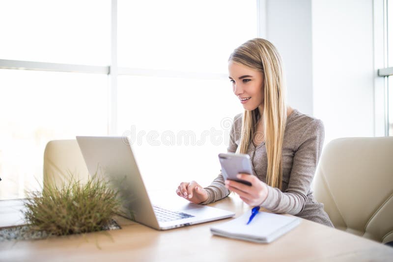 Young business woman with phone in hand using laptop at office