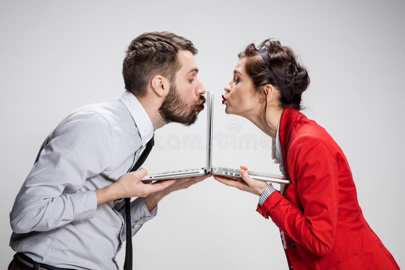 The young businessman and businesswoman with laptops kissing screens on gray background