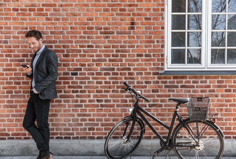 Young businessman going to work on bike commute using his mobile cellphone against city brick wall background. Happy