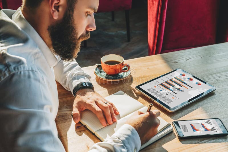 Young businessman is sitting at table and making notes in notebook. On table is tablet computer,smartphone with graphs royalty free stock photos