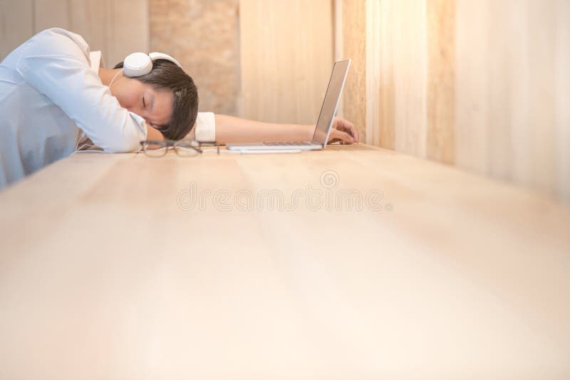 Young Asian businessman take a nap on wooden desk in workspace, casual creative designer resting in studio, tired from overtime work and sleeping in front of laptop computer. Young Asian businessman take a nap on wooden desk in workspace, casual creative designer resting in studio, tired from overtime work and sleeping in front of laptop computer