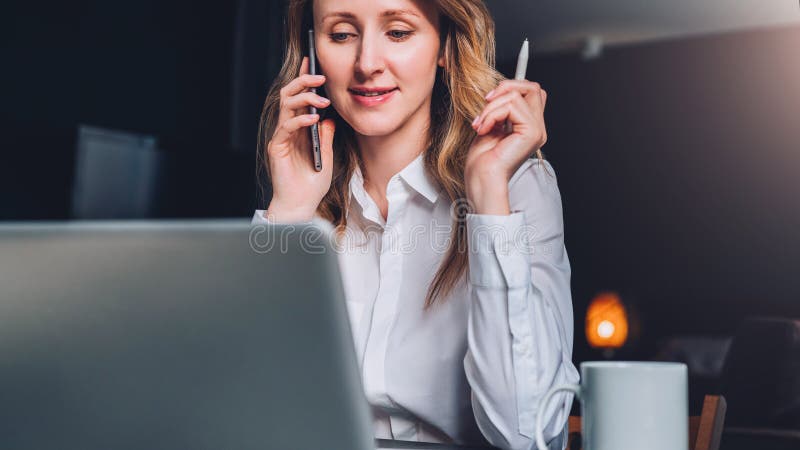 Young businesswoman in shirt is sitting in office at table in front of computer, talking on cell phone