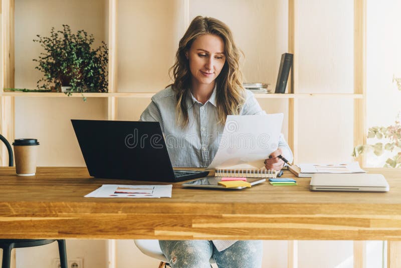 Young businesswoman woman is sitting at kitchen table, reading documents,uses laptop,working, studying.